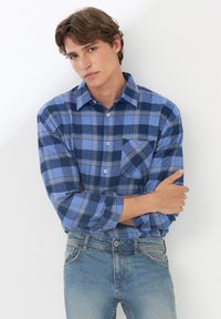 Camicia a quadri blu in un tessuto morbido con maniche lunghe, chiusura frontale con bottoni e taschino sul petto. Indossata con jeans in denim blu chiaro.