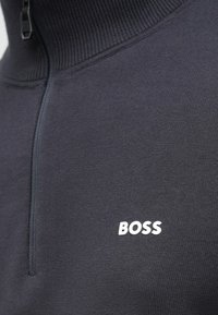 Sweatshirt cinzento escuro com fecho éclair, gola alta, textura canelada e logótipo branco "BOSS" no peito. Confeção em tecido liso.