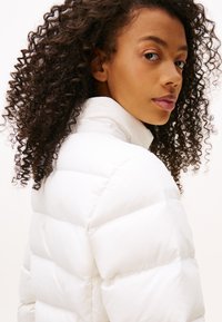 Junge Frau mit lockigem Haar, die über die Schulter schaut, trägt eine weiße Steppjacke mit hohem Kragen.