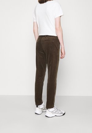 Osoba ubrana w ciemnobrązowe sztruksowe spodnie, białą koszulkę i białe trampki, stojąca tyłem na tle jednolitego jasnego tła.