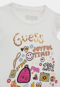 Bela organskega bombaža majica s kratkimi rokavi, ki vsebuje bleščice z napisom "Guess", pisano vezenje grafike, naborke na rokavih in igrive vzorce.