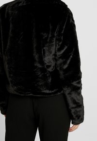 Person wearing a black plush faux fur jacket and black pants, shown from the back against a plain light background.