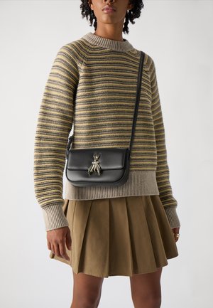 Černá kožená crossbody taška se stříbrným kouzlem v podobě hmyzu, spárovaná se šedým a žlutým pruhovaným pleteným svetrem a béžovou skládací sukní.