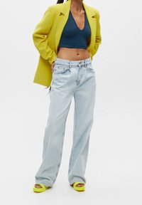 Femme portant un blazer jaune, un top court bleu foncé, un jean large bleu clair et des sandales à plateforme jaunes, debout sur un fond blanc.