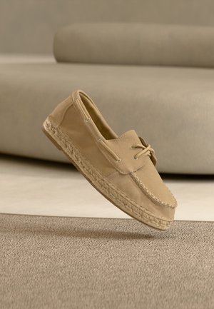 Mocasini bej din piele intoarsă, cu talpă împletită și detaliu cu șiret, plutind deasupra unei mochete bej asortate, cu un fundal neclar al unei canapele.