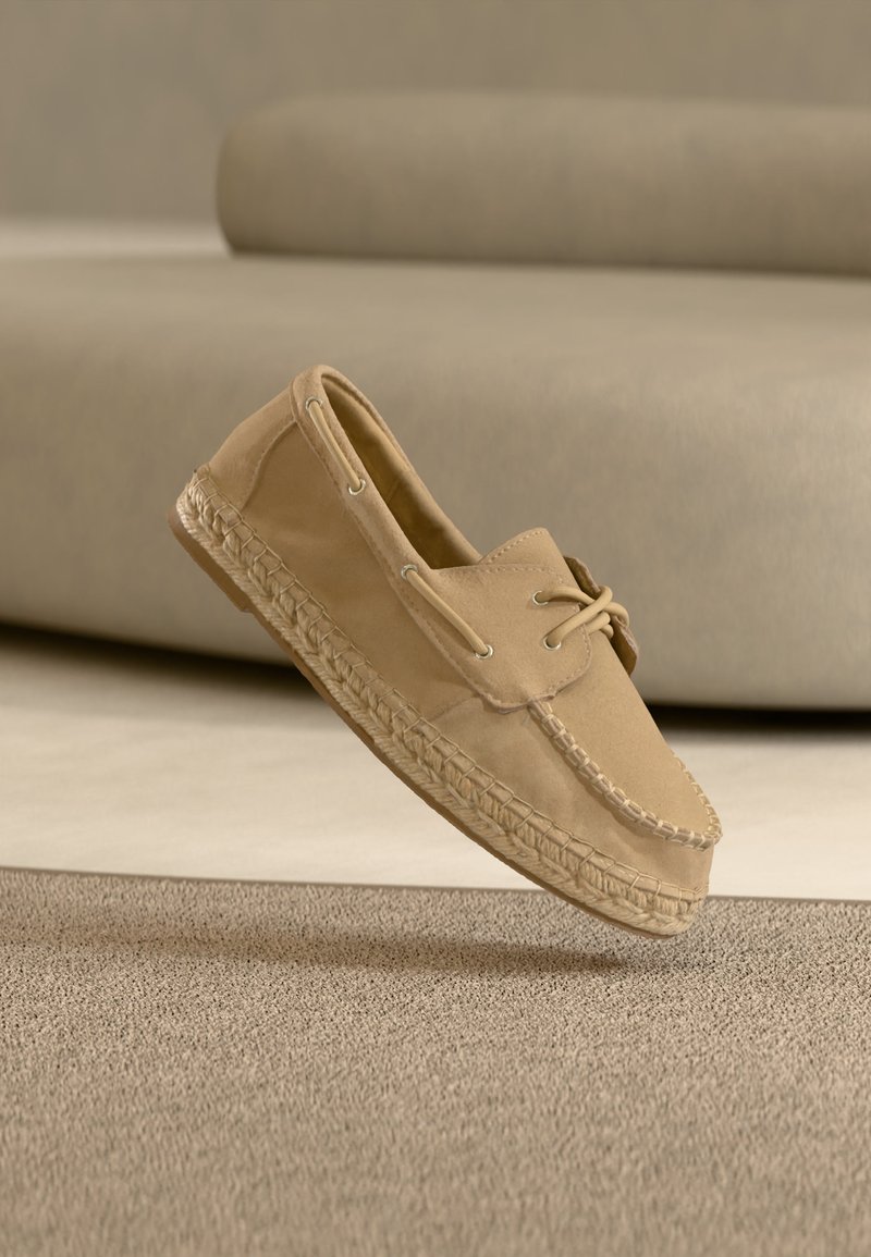 Μπεζ μοκασίνι από σουέντ με πλεκτή σόλα και λεπτομέρεια κορδονιού, αιωρούμενο πάνω από μπεζ χαλί που ταιριάζει, με θολό φόντο καναπέ.