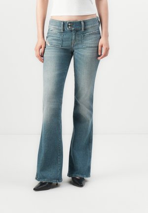 Femme portant un jean flare bleu délavé avec des poches plaquées à l'avant et des talons noirs à bout pointu, debout devant un fond blanc.