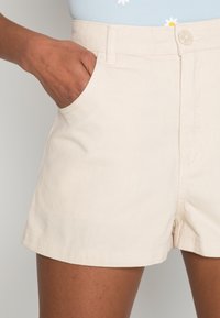 Calções de algodão beije com cintura elástica, bolsos frontais e fecho com botão. A textura é suave com um padrão ligeiramente canelado.