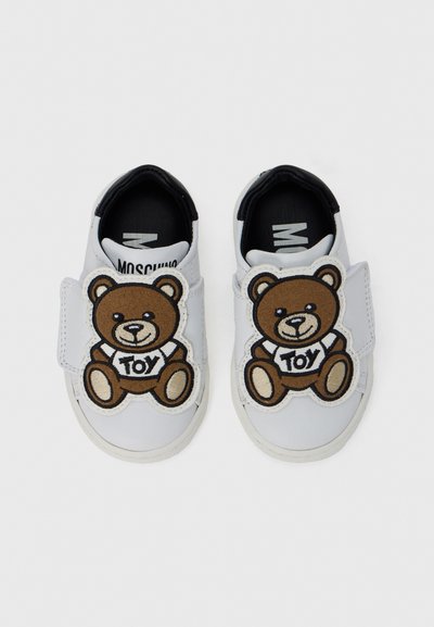Λευκά αθλητικά παπούτσια με ένα καφέ σχέδιο αρκουδάκι teddy και το κείμενο "TOY" στο μπροστινό μέρος. Βέλκρο ιμάντας, λαστιχένια σόλα και μαύρη εσωτερική επένδυση.