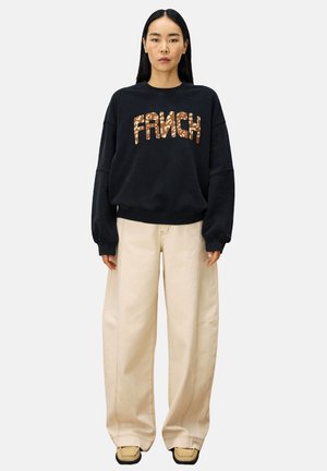 Femme portant un sweat-shirt noir avec le texte "FRNCH" en imprimé léopard, un pantalon beige ample et des chaussures à bout carré beiges, se tenant contre un fond blanc.