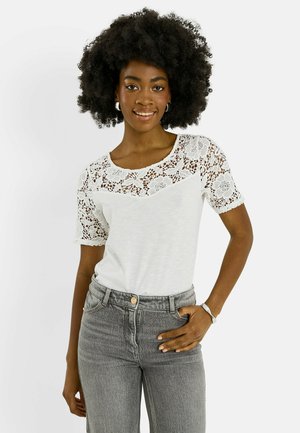 Mujer sonriente con cabello rizado natural que lleva una camiseta corta blanca de encaje floral y vaqueros grises de cintura alta, mano en el bolsillo.