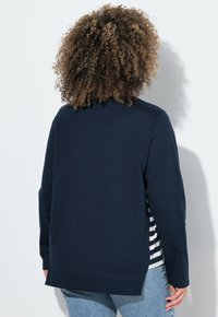 Marineblauer Pullover mit ribbelten Bündchen und Saum, über einem gestreiften Hemd getragen, das am unteren Rand sichtbar ist.