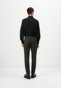 Bărbat stând cu fața la un perete, îmbrăcat cu o cămașă neagră cu mânecă lungă, pantaloni de culoare verde măslin și încălțări cu talpă groasă negre.