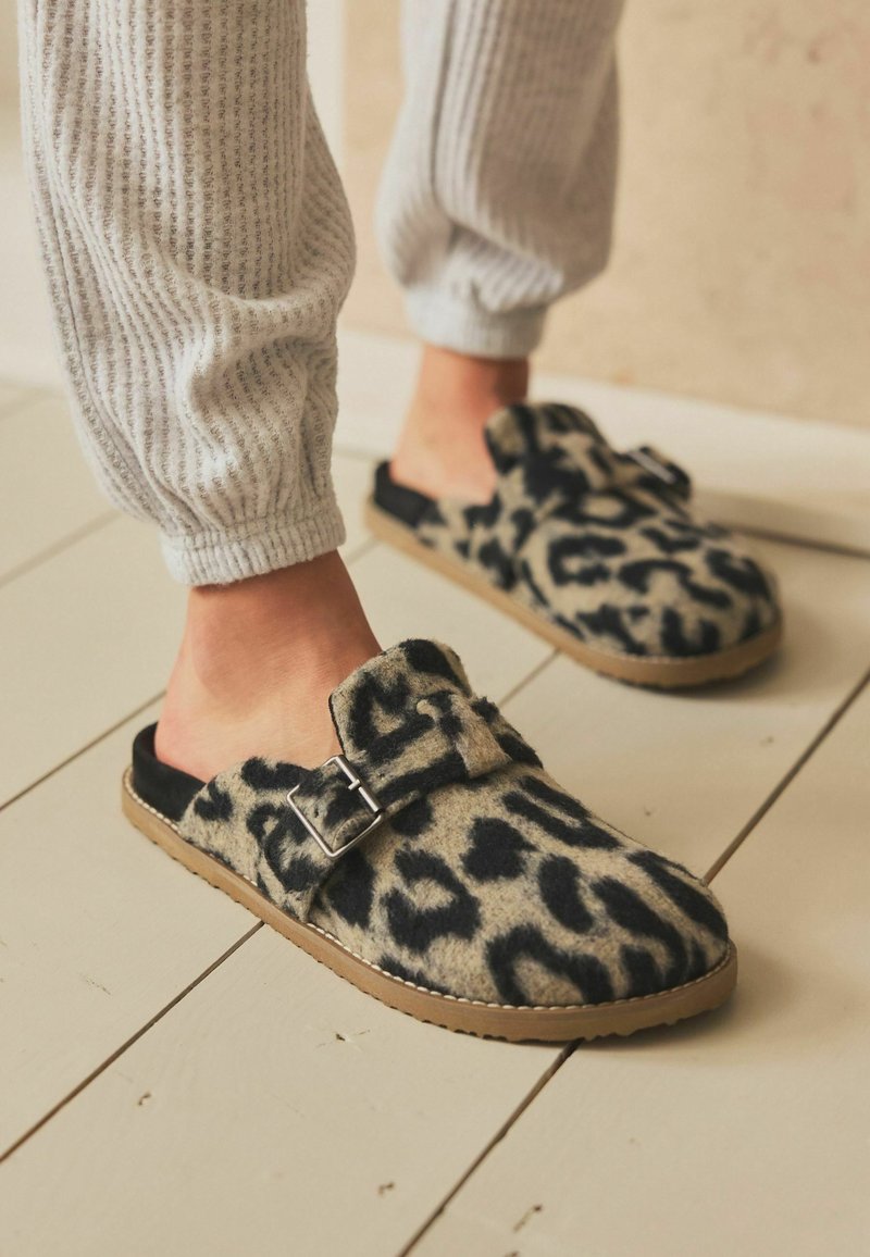 Des pieds portant des sabots en fourrure à imprimé léopard avec des boucles, associés à un pantalon de survêtement gris clair texturé sur un sol en bois.