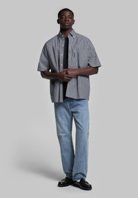 Camicia a maniche corte a righe bianche e nere con colletto a bottoni, indossata sopra una maglietta nera, abbinata a jeans azzurri chiari e scarpe nere.