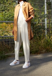 Brun skjorta med knappar över en vit polotröja, i kombination med krämfärgade byxor och vita sneakers med orange ränder.