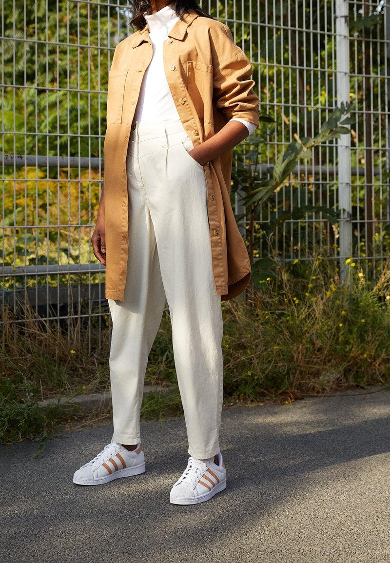 Brun skjorta med knappar över en vit polotröja, i kombination med krämfärgade byxor och vita sneakers med orange ränder.