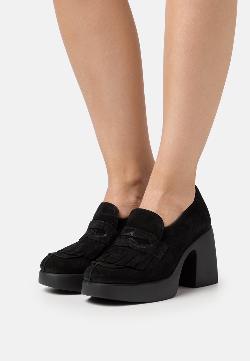 Mocassins à plateforme en daim noir avec détails à franges, portés aux pieds, dotés de talons hauts épais sur un fond clair uni.