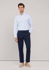 Camicia azzurra a righe bianche con bottoni, pantaloni blu navy, scarpe beige senza lacci, con dettagli del logo discreti sulla tasca della camicia.