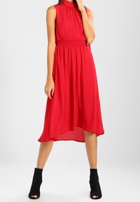 Rode mouwloze jurk met een hoge hals, een gerimpelde taille, een vloeiende rok en een zoom tot op de knie, gecombineerd met zwarte enkellaarsjes.