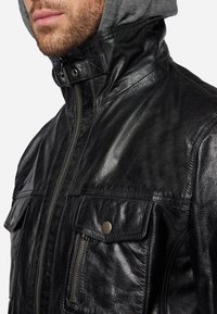 Schwarze Lederjacke mit hohem Kragen, Reißverschluss, zwei Vorderaschen und strukturiertem Finish. Unter dem Kragen befindet sich eine graue Kapuze.