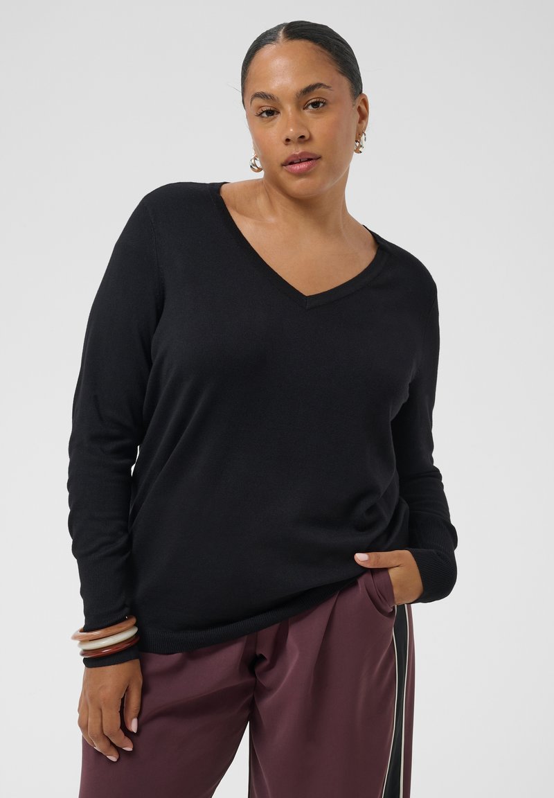Femme aux cheveux plaqués en arrière, portant un pull noir en V, un pantalon bordeaux à bandes latérales, des boucles d'oreilles créoles et des bracelets, posant avec la main dans la poche.