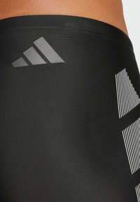 Crne atletske kratke hlače izrađene od glatkog, rastezljivog materijala, s diskretnim sivo Adidas logotipom i bijelim vertikalnim prugama sa strane.