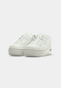 Baskets en cuir blanc avec une texture lisse, bout arrondi, semelle plateforme épaisse et perforations discrètes. Comprend des lacets et des détails de logo.