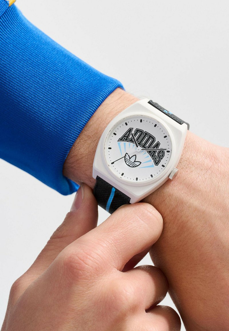 Montre analogique blanche avec le logo Adidas, bracelet texturé noir avec des accents bleus, cadran rond et indiqueurs d'heures et de minutes simples.