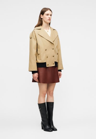 Γυναίκα όρθια, φοράει μπεζ διπλόσαρκο σακάκι, καφέ δερμάτινη φούστα και μαύρες ψηλές μπότες με τακούνι ως το γόνατο, σε απλό φόντο.