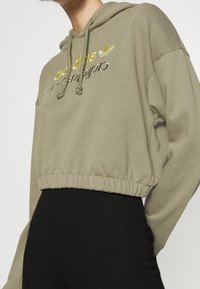 Person wearing cropped olive green hoodie with yellow and white Adidas logo and black high-waisted pants against white background.