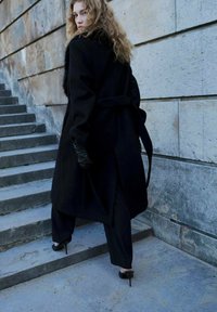 Abrigo de lana negro con cuello de piel, cinturilla ceñida y mangas largas, combinado con pantalones negros ajustados y zapatos de tacón alto; de pie en unas escaleras.