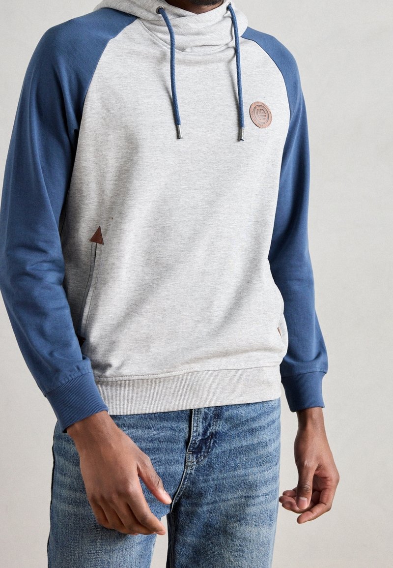 Homme portant un sweat à capuche gris clair avec manches bleues et cordons bleus, associé à un jean bleu, debout devant un fond uni.