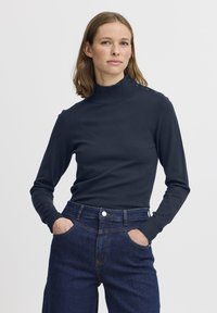 Maglione a collo alto blu navy con maniche lunghe e dettagli con bottoni sulle spalle, abbinato a jeans scuri a vita alta.