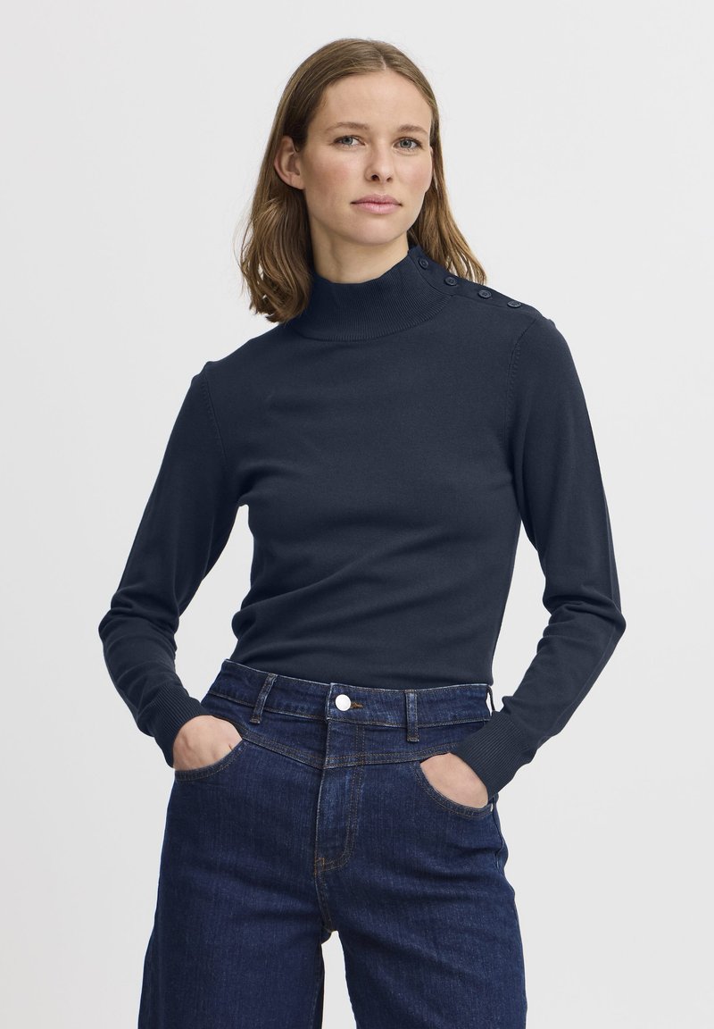 Maglione a collo alto blu navy con maniche lunghe e dettagli con bottoni sulle spalle, abbinato a jeans scuri a vita alta.