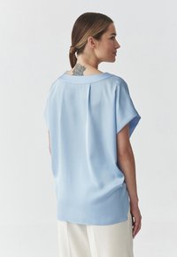 Blusa azzurra chiara a maniche corte con vestibilità rilassata, scollo rotondo e piega sulla schiena. Tessuto morbido con un leggero luccichio.
