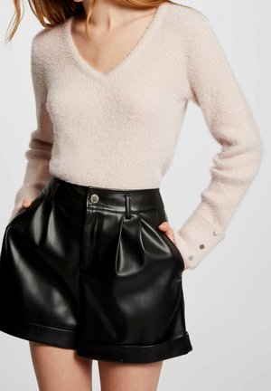 Femme portant un pull beige doux à col en V et un short noir plissé en cuir synthétique taille haute, les mains dans les poches.