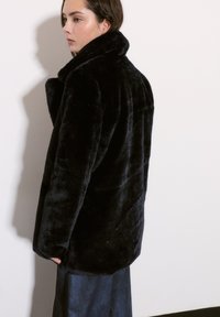 Czarny futrzany płaszcz z wysokim kołnierzem, luźny krój i gładka faktura, noszony na ciemnoniebieskiej spódnicy, na jednolitym tle.