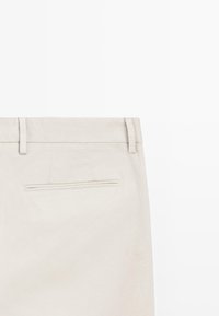 Beiga byxor som visar bakre midjeband med bältesöglor och en enkel fuskficka mot en enfärgad vit bakgrund.