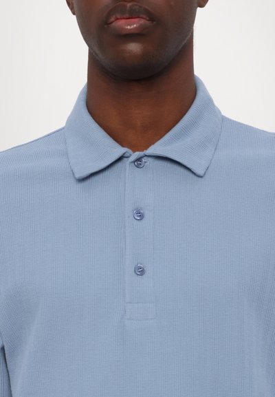Šviesiai mėlynas polo marškinėlis, pagamintas iš tekstūruotos medžiagos, su apykakle ir trimis mygtukais. Mygtukai apvalūs ir suspausti kraštuose.