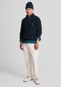 Jersey de punto azul marino con cuello alto simulado y cremallera de cuarto de vuelta, superpuesto sobre una camisa verde, combinado con pantalones crema y zapatillas de color claro.