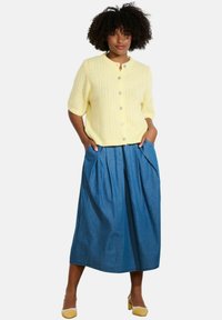 Gaišdzeltena, īsām piedurknēm adīta jakas ar pogām, apvienota ar garu, krokotu džinsu svārku un dzeltenām papēžu kurpēm.