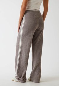 Szare welurowe spodnie dresowe z białymi paskami po bokach, szerokie nogawki, miękka tekstura i elastyczny pas. Widoczny detal szwu na dolnym końcu.