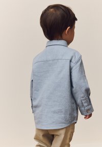 Camicia celeste chiaro con bottoni, realizzata in tessuto morbido e strutturato. Presenta un colletto, maniche lunghe e un orlo curvo. Indossata con pantaloni beige.