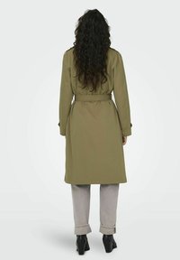 Olivgrön omslagskappa med bälte, långa ärmar och rak passform, kombinerad med grå jeans med uppvikta benslut och svarta klackstövlar.