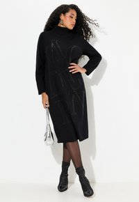 Abito maglione nero con scollatura alta e maniche lunghe, decorato con scritte metalliche. Abbinato a stivaletti neri e una borsa argentata.