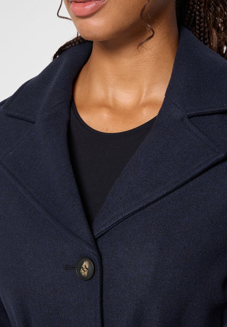 Manteau en mélange de laine bleu marine avec un col cranté, doté d'une fermeture à un bouton et d'une texture douce. Superposé sur un haut noir.