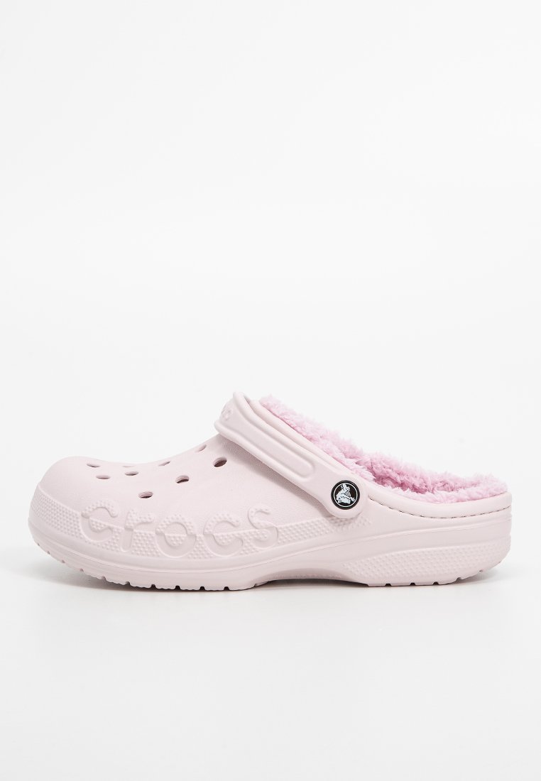 Light pink clogs with a fuzzy inner lining, vent holes on the sides, and a black circular logo on the strap. Made of rubber-like material.