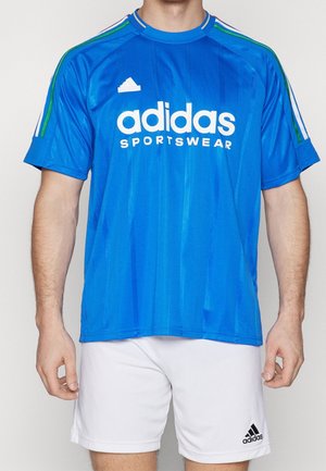 Mężczyzna noszący niebieską koszulkę sportową Adidas z białymi i zielonymi paskami na rękawach oraz białe spodenki Adidas.