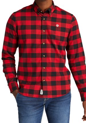 Mann trägt ein rot-schwarz kariertes Hemd mit langen Ärmeln und blaue Jeans, Hände in den Taschen, Oberkörper sichtbar.